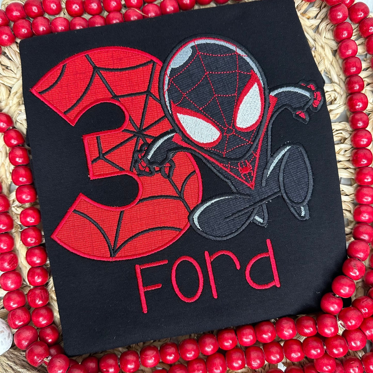 An inspired miles spider man design is appliqued in black fabric on a black shirt along with a custom birthday number in red fabric with black thread to make an intricate web design. Personalized with a name in red thread. 