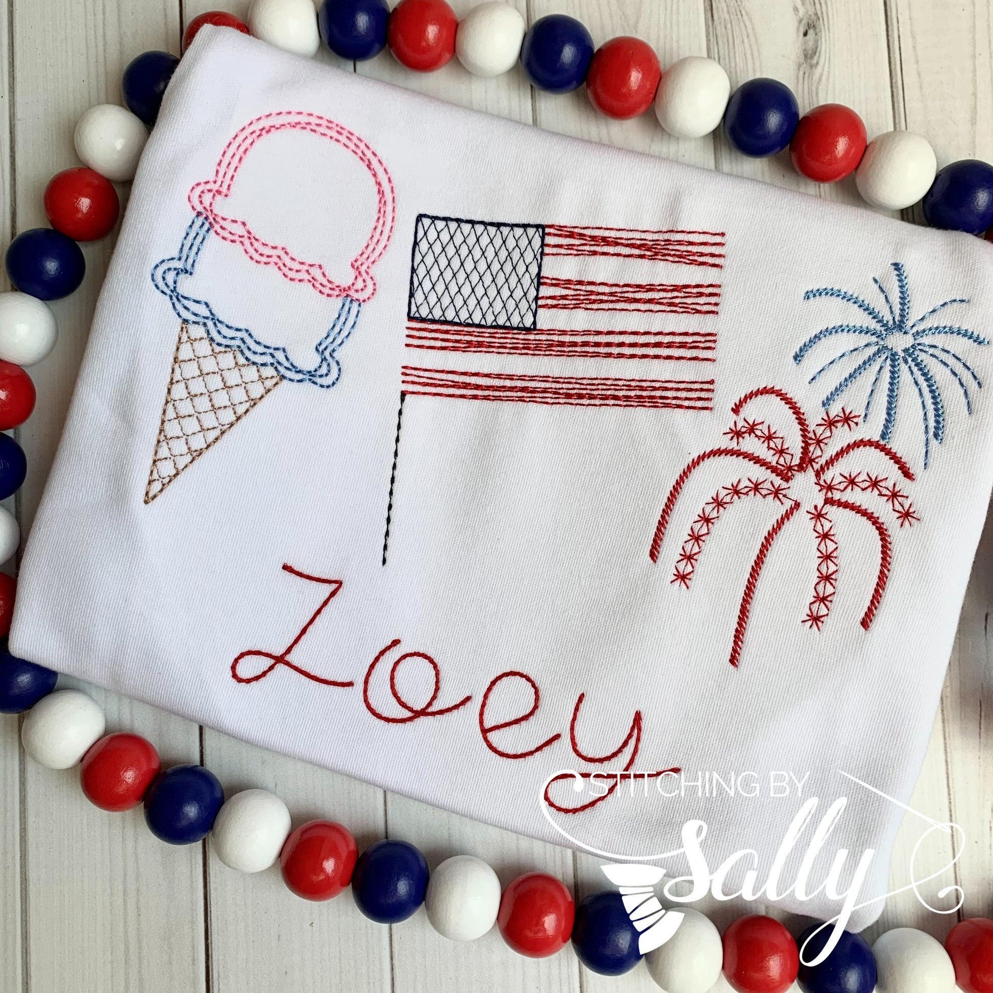 A sketch design of ice cream, American flag, and fireworks are embroidered onto a white shirt with a name underneath. 