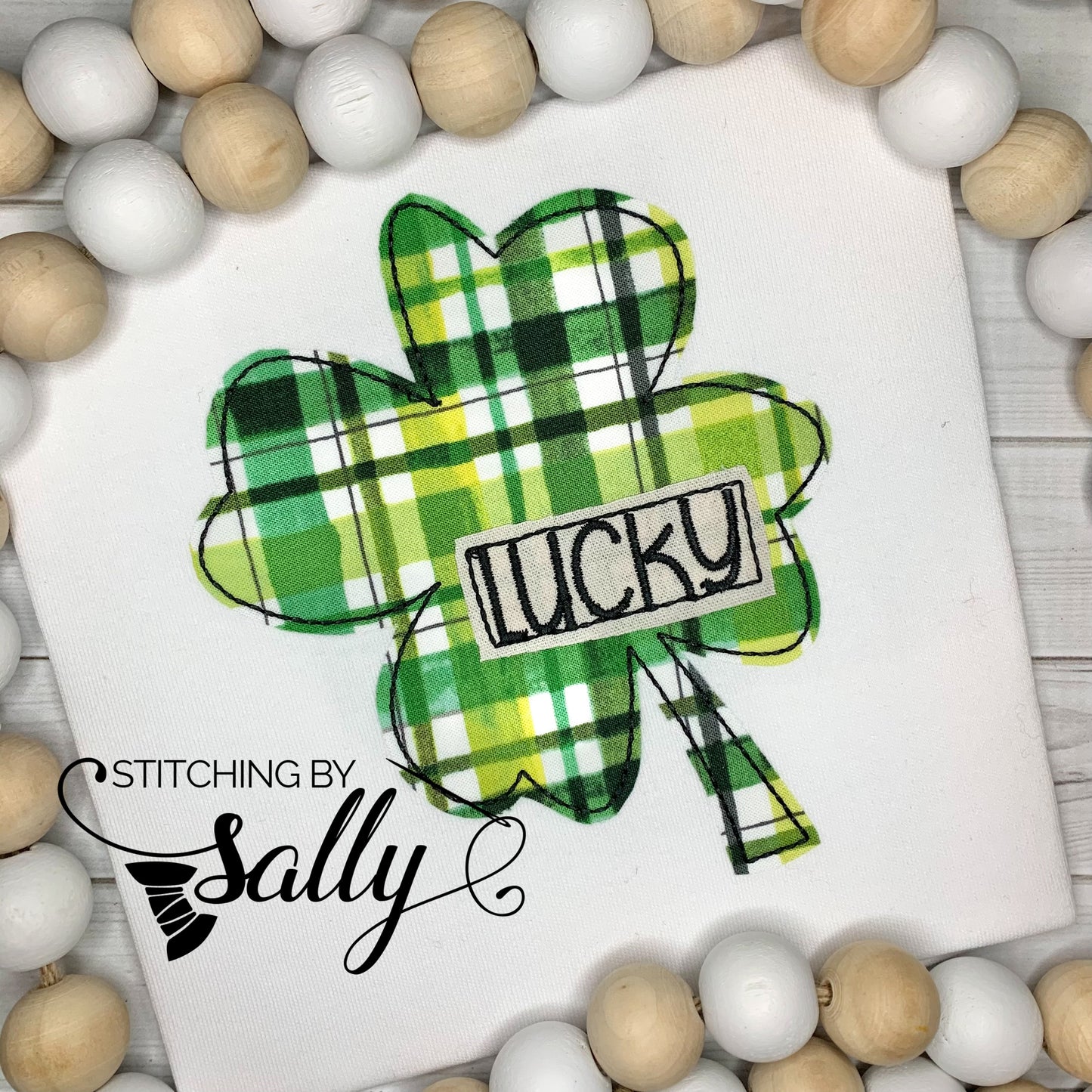 A white shirt with a 4 leaf clover appliqued in green plaid pint with lucky embroidered on the clover.