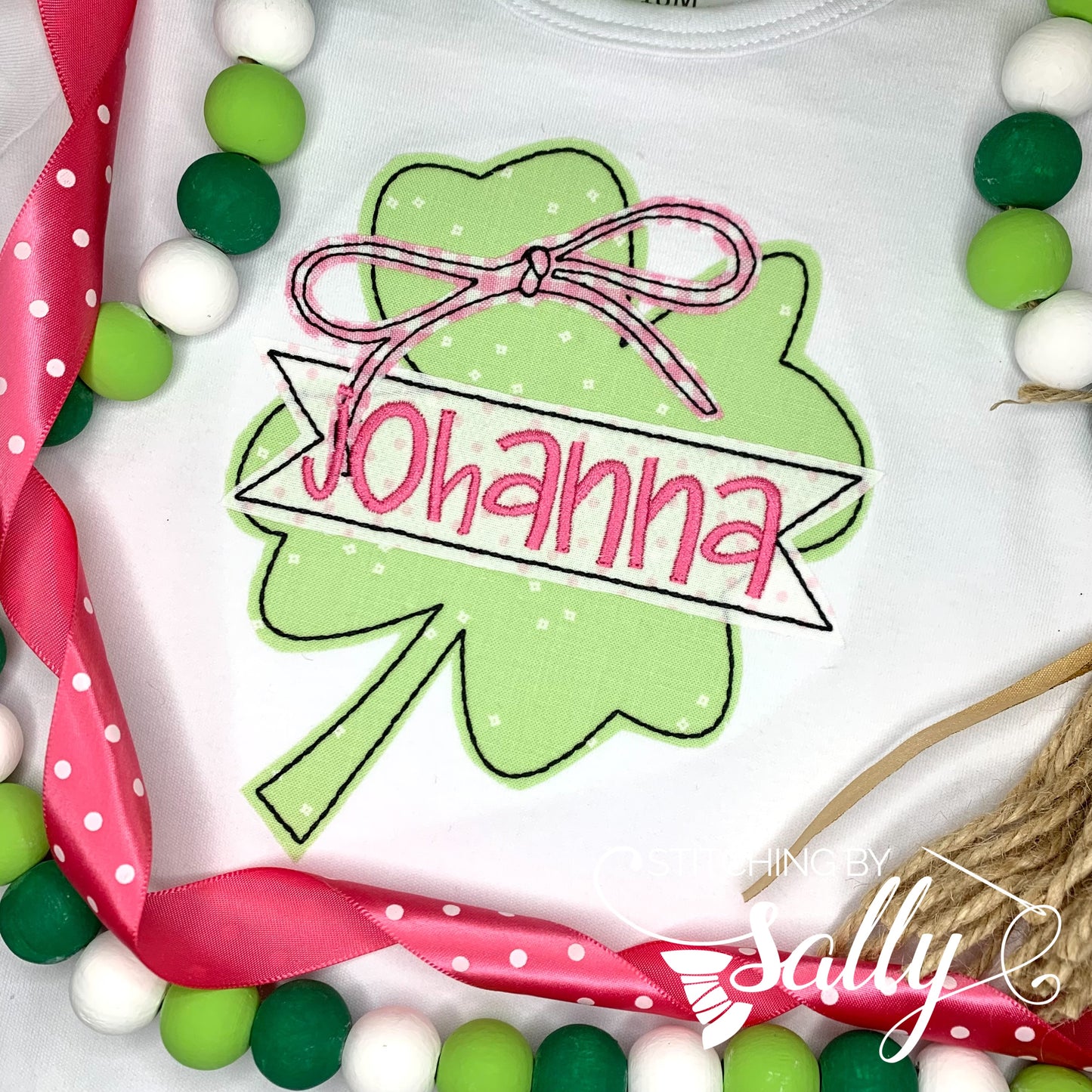 A white shirt featuring a clover applique with a bow in pink gingham fabric on top. A name is embroidered across the clover. 