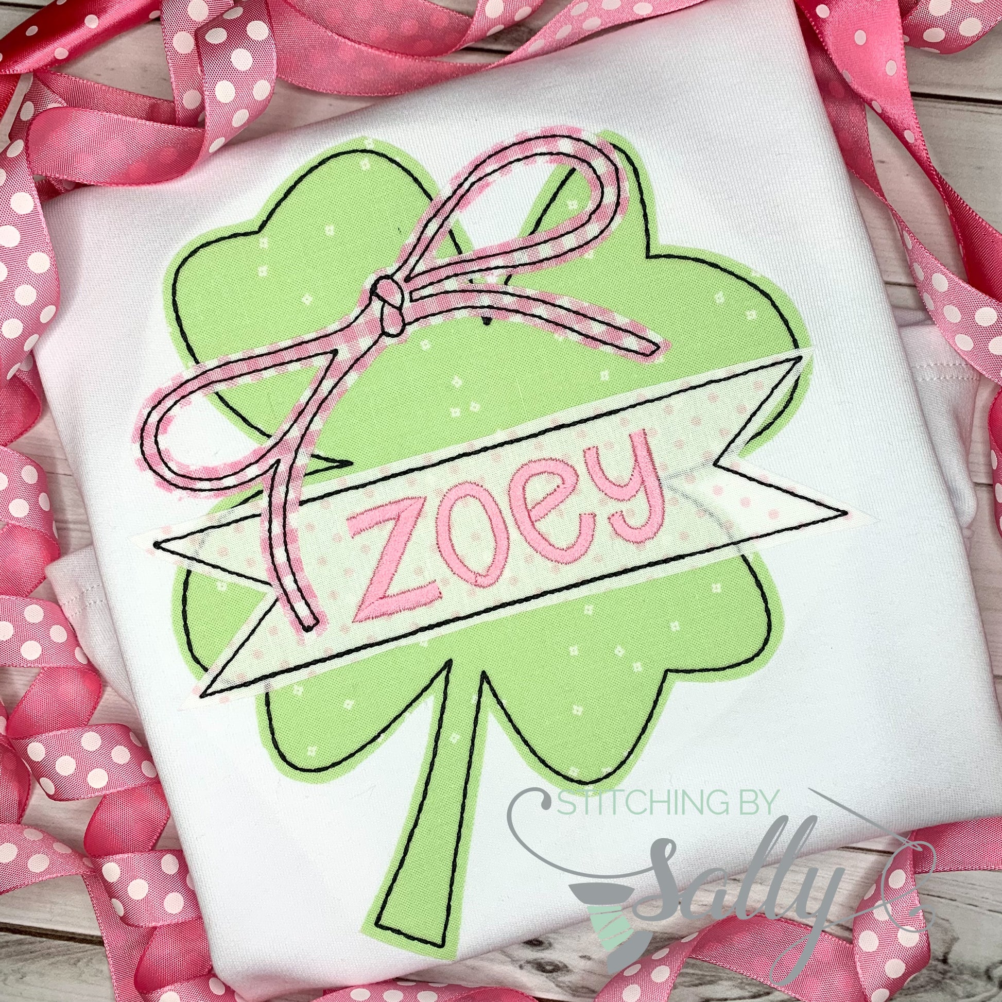 A white shirt featuring a clover applique design with a pink gingham bow. A name is embroidered across the clover in pink thread. 