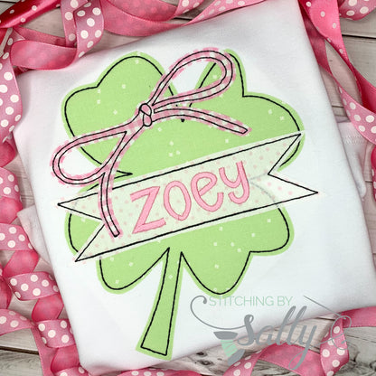 A white shirt featuring a clover applique design with a pink gingham bow. A name is embroidered across the clover in pink thread. 