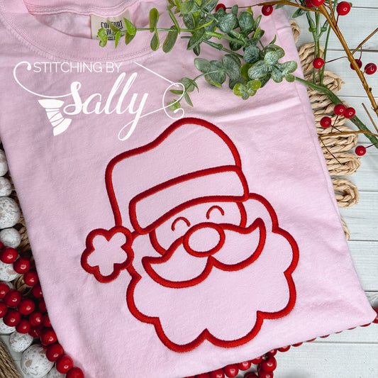 A pink shirt with a jolly Santa face embroidered in red thread on the front center of the shirt. Stitching by Sally 