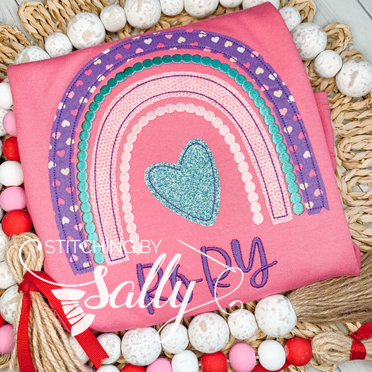 A pink shirt with a purple, pink and teal rainbow appliqué design. A heart and name are embroidered underneath. 