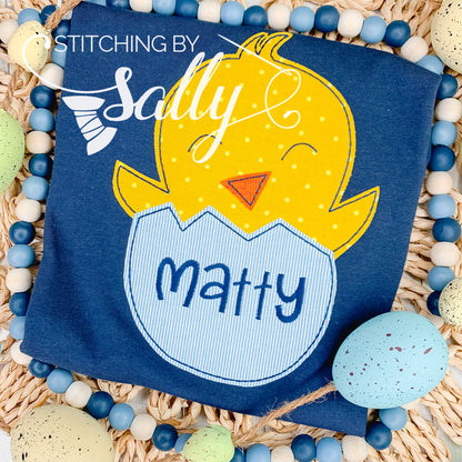 A cute Easter chick popping out of an egg is appliqué on to a blue shirt. A name is embroidered on the egg. 