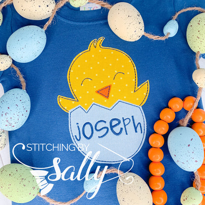 A design of baby chick hatching from an egg is appliquéd on a blue shirt. A name is embroidered on egg. 