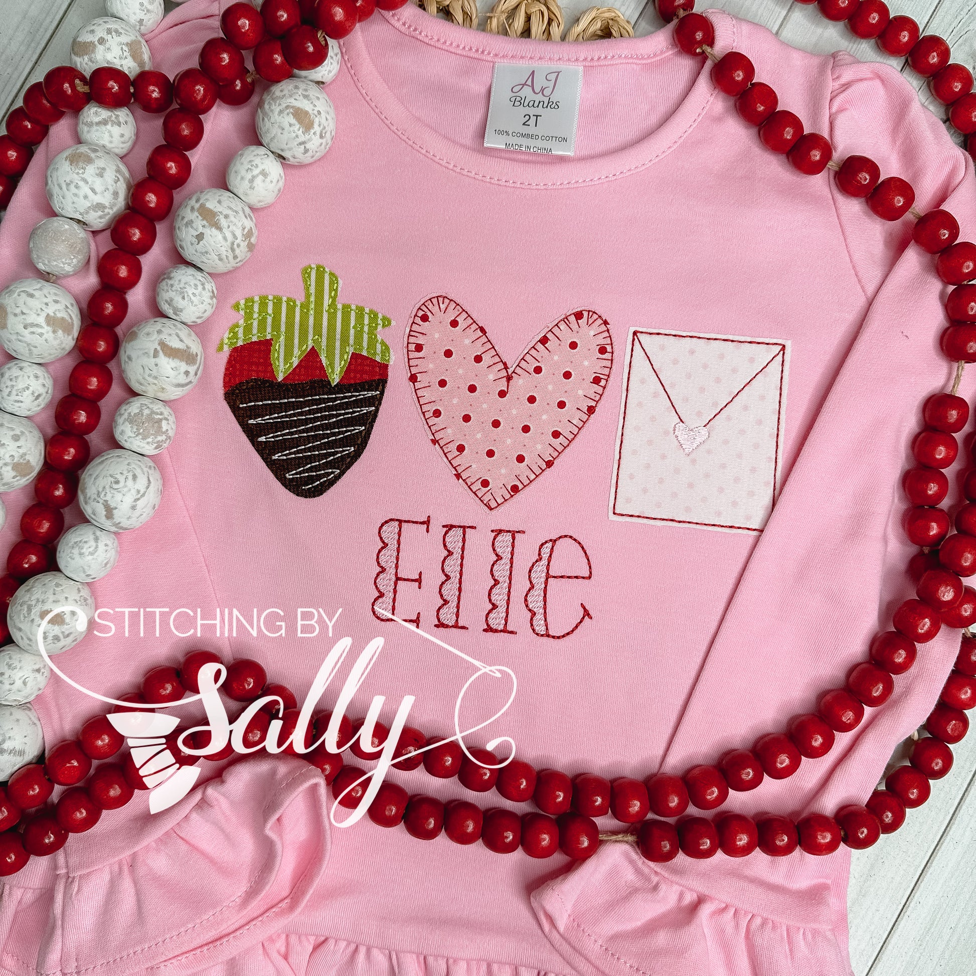 A pink shirt with a strawberry, heart and letter appliqué design and an embroidered name below.