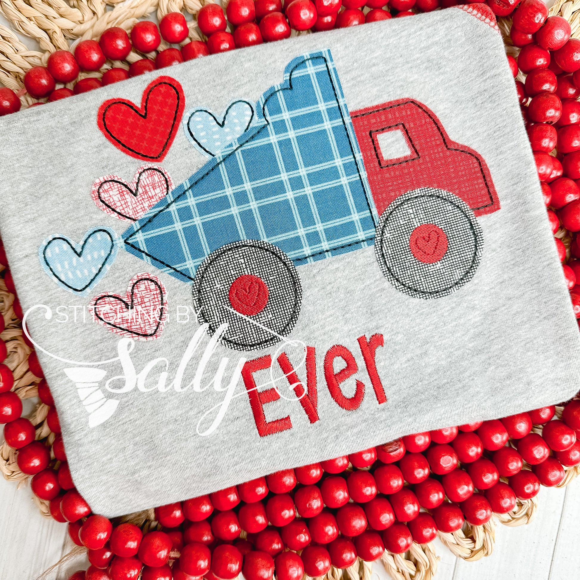 A grey shirt with a red and blue dump truck appliqué design. Truck is dumping red heart appliqués. 