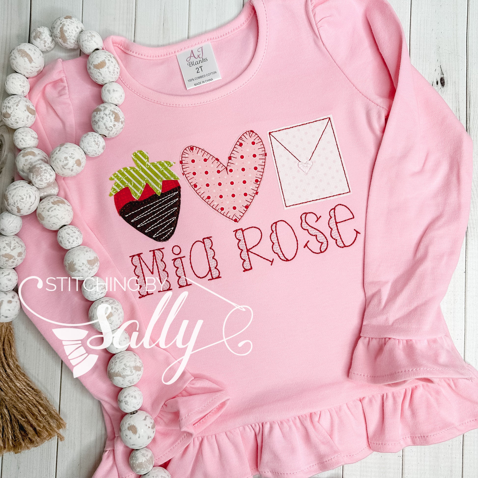 A strawberry, heart, and letter appliqué on a pink shirt with a name embroidered underneath.