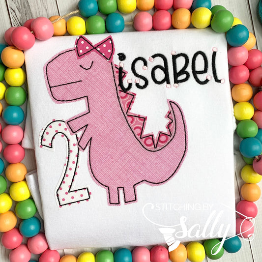 A cute pink dinosaur is appliquéd onto a white shirt. The dinosaur applique has a pink bow and is holding a number 2 in pink polka dot fabric. a name is embroidered in black thread.