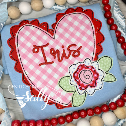 A blue shirt with heart appliquéd in pink gingham fabric with red fabric ruffles. A cute pink and red flower is off the side of the heart. A name is embroidered in red thread.  