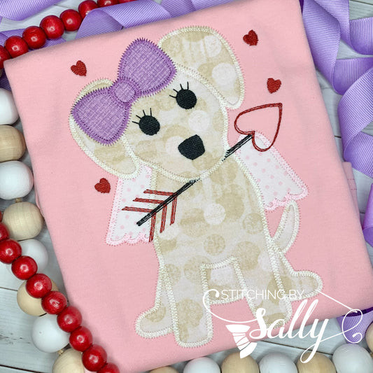 A pink shirt with a Cupid dog appliqué in tan fabric with a purple bow. The dog is holding an arrow in its mouth and a few red hearts are embroidered around. 