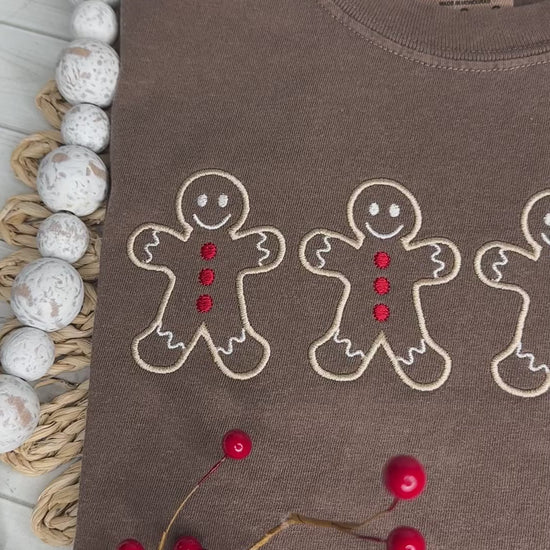 A video of a brown shirt with four Gingerbread men embroidered in tan thread with red buttons. Stitching by Sally 